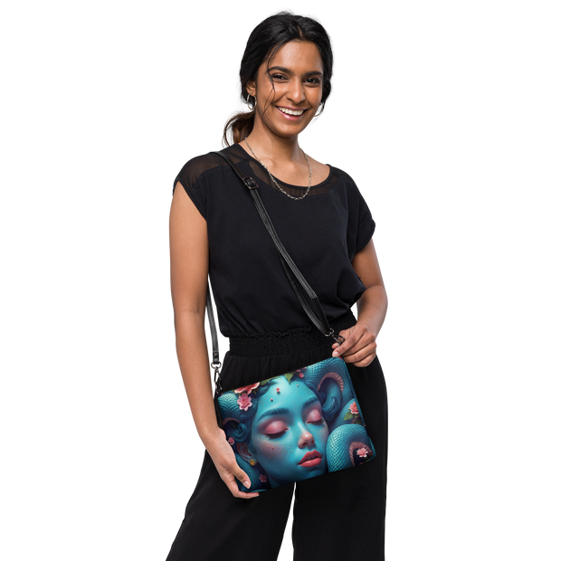 A crossbody bag featuring a vibrant, surreal graphic print of a sleeping woman's face with blue snakes and pink flowers wrapping around her, complete with a black faux-leather wrist and shoulder strap.