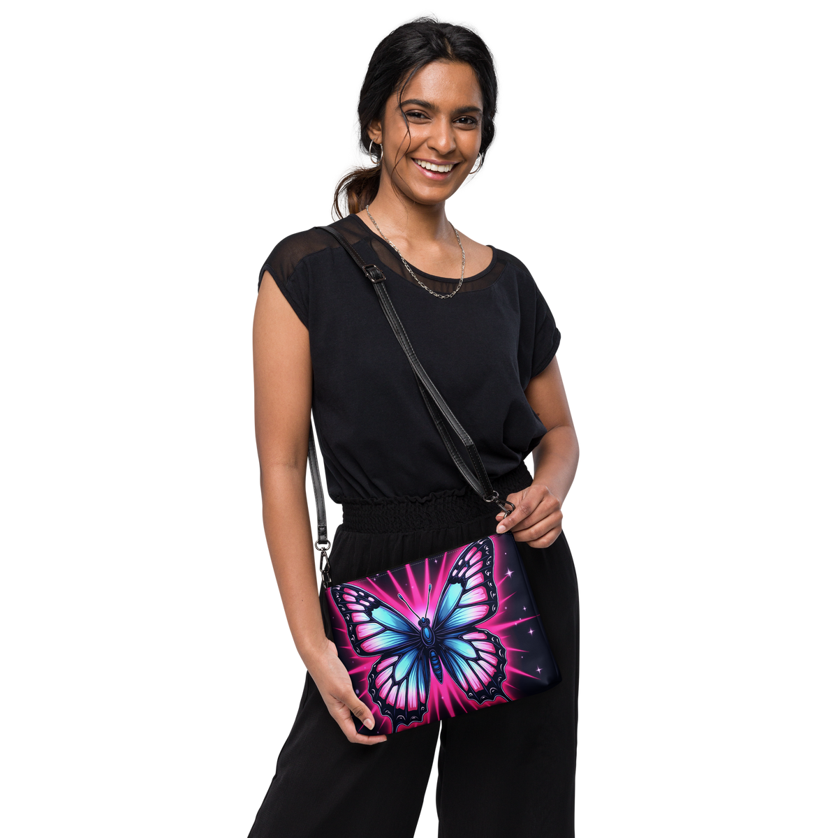 A woman wearing a black top and black pants models a black crossbody bag featuring a vibrant, colorful butterfly print with pink, blue, and purple hues on a black background.