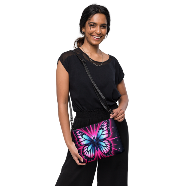 A woman wearing a black top and black pants models a black crossbody bag featuring a vibrant, colorful butterfly print with pink, blue, and purple hues on a black background.