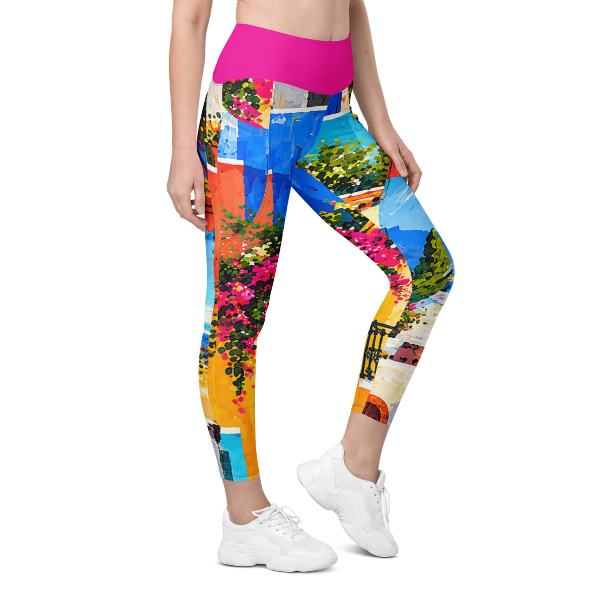 A model is wearing colorful, high-waisted leggings with a vibrant Mediterranean-style print featuring blue sky, orange buildings, and pink flowers. The leggings have a solid bright pink waistband and are styled with white sneakers against a plain white background.
