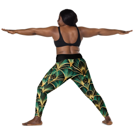 A woman poses in a yoga warrior II stance wearing high-waisted leggings with a green and gold palm leaf pattern, paired with a black sports bra, against a solid white background.