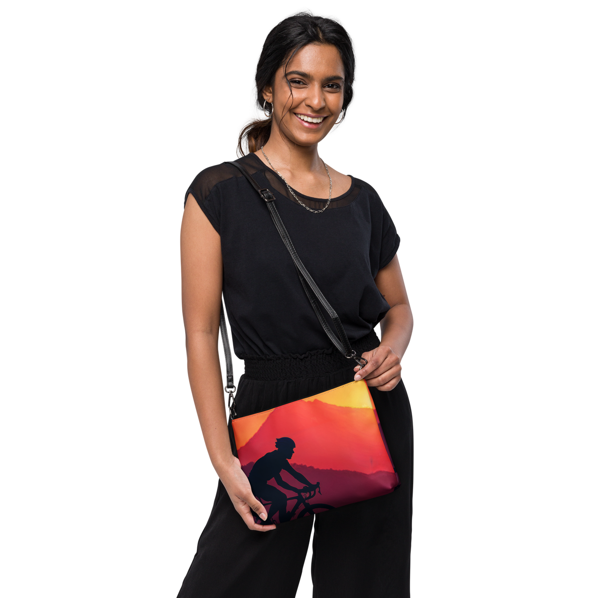 A woman wearing a black top and black pants models a black crossbody bag with a vibrant sunset print featuring a cyclist silhouette. The bag has a black strap and is shown against a solid white background.